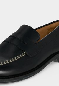 Mocassin en cuir noir avec une couture blanche contrastante le long de la pointe et une large bride sur le dessus, présenté sur un fond uni.
