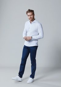 Witte, langmouw poloshirt, blauwe broek en witte sneakers. Eenvoudig ontwerp, gladde textuur en slim fit. Handen voor zich gevouwen.