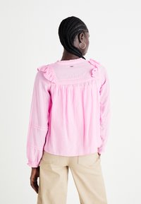 Blusa a maniche lunghe rosa chiaro con dettagli di rouches sulle spalle, schiena arricciata e polsini elasticizzati. Tessuto morbido, vestibilità casual.