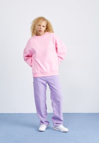 Persoană cu păr blond creț, purtând un pulover roz supradimensionat, pantaloni lila și adidași albi, stând pe un podea albastră împotriva unei pereți albe.