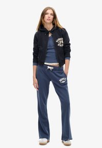 Marineblå hættebluse med snøre og logo, parret med løse marineblå bukser med hvidt print. Stoffet fremstår blødt og afslappet. Hvide sneakers.