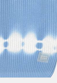 Coperta lavorata a maglia di colore azzurro chiaro con motivi a nuvola bianchi. Presenta bordi a coste e un'etichetta con la scritta "SCOTCH & SODA AMSTERDAM."
