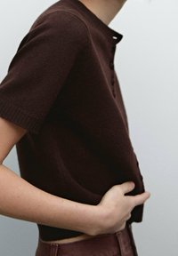 Osoba w ciemnoszarym, krótkoz rękawowym swetrze z zapięciem na guziki z przodu, z jedną ręką opartą na biodrze na jednolitym jasnym tle.