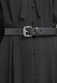 Ceinture en cuir noir avec une boucle en argent, passée à travers des trous, portée sur un vêtement en tissu noir avec des détails de boutons. Texture lisse.