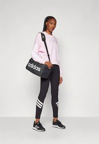 Mulher com uma sweatshirt rosa da Adidas, leggings pretas com riscas brancas, sapatilhas pretas, a carregar uma mala de desporto preta da Adidas ao ombro.
