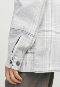 Camisa de cuadro gris con líneas blancas y negras, textura suave, puños abotonados y aberturas laterales para facilitar el movimiento.