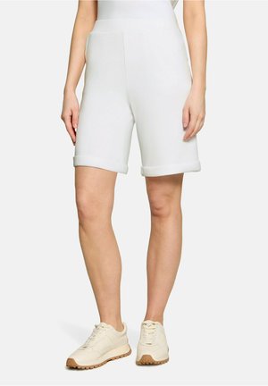 Personne portant un short blanc mi-genou avec des ourlets roulés et des baskets beiges, debout devant un fond clair uni.
