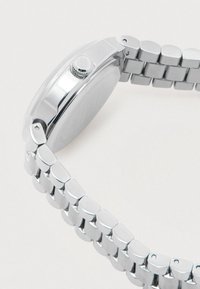 Silver metal watch band with linked segments attached to a round, polished watch case viewed from the side against a white background.