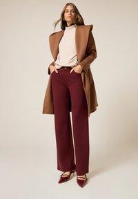 Bruin wollen jas, beige coltrui en bordeaux brede broek. Gespitste bordeaux hakken met banden. Neutrale achtergrond.