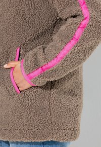 Veste en polaire marron avec une surface texturée, comportant des bandes d'accent rose le long des manches et une poche latérale.