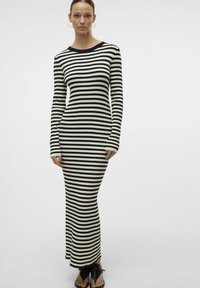 Robe longue côtelée présentant des rayures horizontales noir et blanc, un col rond et une silhouette ajustée. Portée avec des sandales noires à bout ouvert.