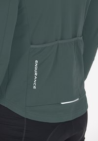 Dark green technical long-sleeve shirt with a textured surface, featuring a rear pocket and reflective accents. Brand logo "ENDURANCE" on the side.