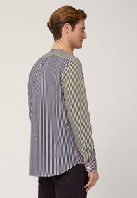 Camicia a righe con diversi colori: blu navy, verde e bianco. Colletto a righe arancione e bianco. Polsini con bottoni, orlo leggermente curvato.