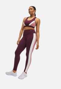 Conjunto de sujetador deportivo y leggings en burdeos, rosa y blanco. Presenta un diseño a bloques de color, textura suave y cintura elástica. Zapatillas deportivas blancas.