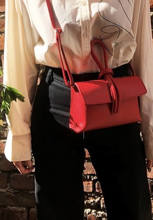 Blouse blanche avec un dessin en ligne noire rentrée dans un pantalon noir, accessoirisée d'un petit sac bandoulière en cuir rouge avec un détail de sangle bouclée.