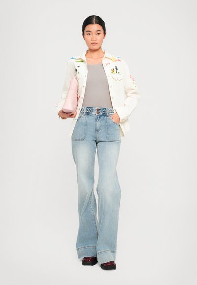Veste blanche à boutons avec broderies colorées, haut gris, jeans évasés bleu clair avec une taille détaillée, tenant un petit sac à main rose.