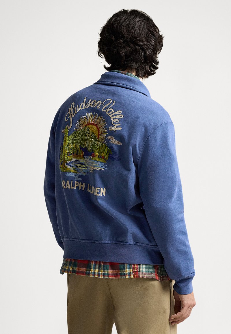 Plava majica s kapuljačom s izvezenim natpisima "Hudson Valley" i "Ralph Lauren" te detaljnim prikazom planine i izlaska sunca. Materijal od runa, opušteni kroj.