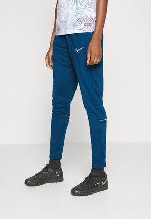 Osoba ubrana w niebieskie spodnie dresowe Nike i czarne buty sportowe Nike stoi na jednolitym jasnym tle.