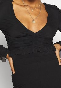 Svart topp med djup V-ringning, rynkdetaljer vid bröstet och volangärmar. Lager av silversmycken kompletterar designen.