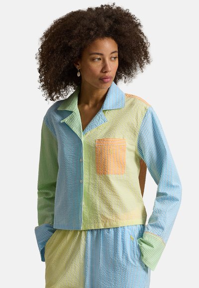 Femme aux cheveux bouclés portant une chemise à manches longues à rayures pastel avec poche avant et pantalon assorti, mains dans les poches, regardant sur le côté.