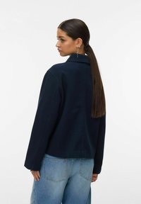 Temno modra skrajšana jakna s ovratnikom, dolgimi rokavi in gladko teksturo. Kombinirana s svetlo modrimi visokim pasom kavbojkami.