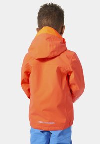 Heldere oranje waterdichte jas met capuchon, voorzien van reflecterende accenten en een logo op de onderrug, gecombineerd met blauwe broek.