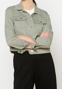 Osoba w jasnozielonej, skróconej kurtce, nosząca białą bluzkę i czarne spodnie, stoi z założonymi rękami na tle jednolitego tła.