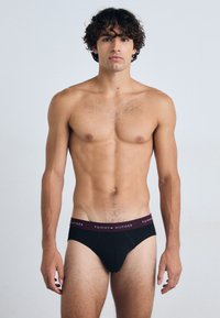 Schwarze Unterhose mit einem breiten bordeauxfarbenen Gummibund, der das Logo "TOMMY HILFIGER" trägt. Leichte Stoffqualität und glatte Textur. Das Modell steht aufrecht.