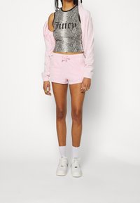 Femme portant un crop top gris motif peau de serpent avec le texte « Juicy », un short rose clair, une veste assortie, des chaussettes blanches et des baskets blanches.