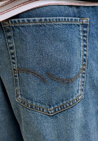 Blauer Denim-Stoff mit sichtbarer Textur. Verfügt über eine aufgenähte Gesäßtasche mit gewölbtem Nahtdesign und kontrastierendem orangefarbenem Faden.