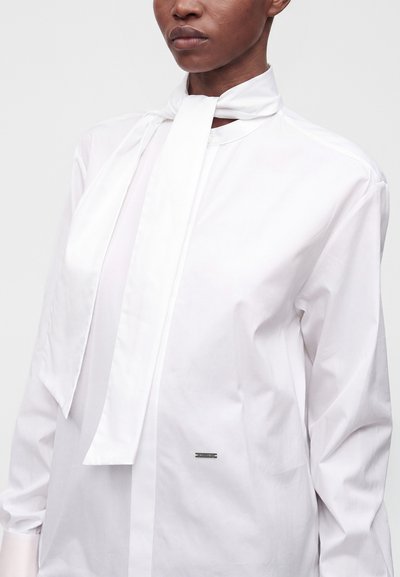Femme portant un chemisier blanc à manches longues avec un foulard noué au cou et un petit logo métallique sur la poitrine.