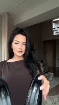 Mulher com cabelo escuro e longo, dentro de casa, segurando e mostrando um par de sapatos de salto altos pretos numa sala de estar moderna.