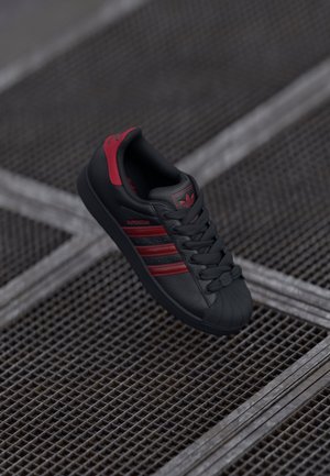 Czarne skórzane sneakersy z czerwonymi akcentami i trzema paskami. Posiadają zaokrąglony nosek oraz teksturowaną gumową podeszwę z sznurowanym designem i logo.