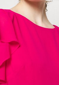 Fuchsia blouse featuring a smooth texture, round neckline, and fluted sleeves. Focused on a particular part of the shoulder and chest area.