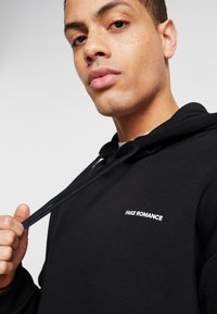 Black hoodie made of cotton with drawstrings. Features "FAKE ROMANCE" embroidered in white on the chest. Smooth texture with a relaxed fit.