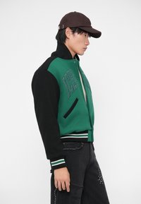 Giacca varsity verde e nera con polsini e vita a costine, caratterizzata da scritte ricamate e tasche laterali, abbinata a un cappellino marrone.