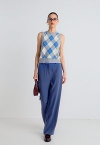 Αμάνικο πουλόβερ ανοιχτού γκρι χρώματος με μπλε σχέδιο argyle, σε συνδυασμό με μπλε παντελόνι φαρδιάς γραμμής. Αξεσουάρ περιλαμβάνουν μια μικρή μπορντό τσάντα χειρός.