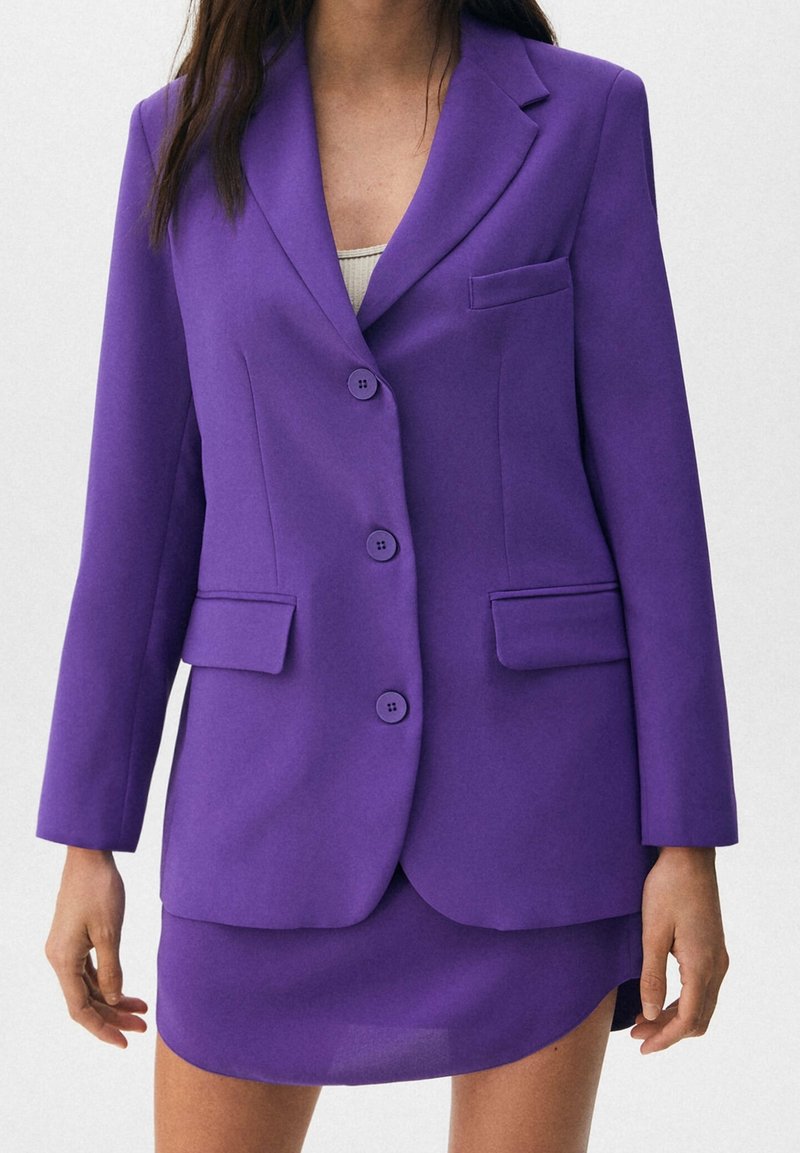 Veste violet en tissu lisse, dotée de revers à crans, de trois boutons et de deux poches avant. Portée avec une jupe assortie.