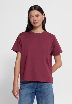 Femme aux longs cheveux foncés portant un t-shirt bordeaux uni à manches courtes et un jean bleu, debout devant un fond uni clair.