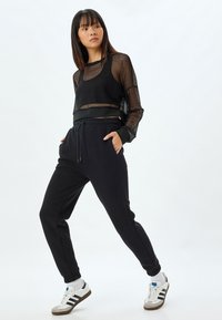 Sort mesh langærmet top over en sort elastisk talje jogger. Modellen har hvide sneakers med sorte striber og grå detaljer på.