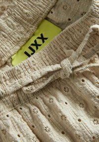 Beige Baumwollshorts mit einem gerafften Bund, dekorativer Stickerei und einer Schleifenbindung sowie einem sichtbaren Etikett in leuchtendem Gelb.