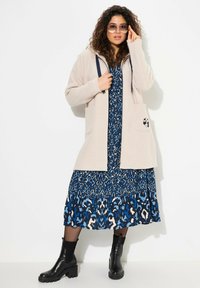 Chaqueta de forro polar beige con detalles en azul marino y bolsillos, superpuesta a un vestido azul con estampado. La modelo lleva botas negras hasta el tobillo y gafas de sol.