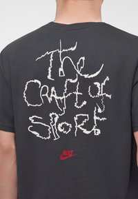 Person wearing a dark t-shirt with white distorted text "The Greatest Story" and a small red Nike logo below on the back.