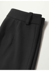 Pantalon noir sur mesure plié, montrant la ceinture avec passants et une couture latérale visible sur fond blanc.