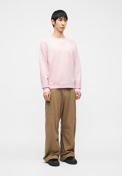 Jeune homme debout portant un sweat-shirt rose clair, un pantalon marron ample avec de grandes poches, et des chaussures noires sur un fond blanc.