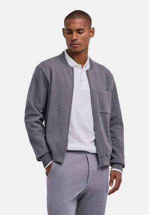 Man wearing grey zip-up jacket over a white polo shirt and matching grey pants, with hands in pockets, looking down.