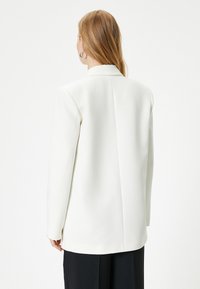 Hvid struktureret blazer med reverskrave, lange ærmer og en ren bagsøm, parret med sorte bukser. Glat tekstur, minimalistisk design.
