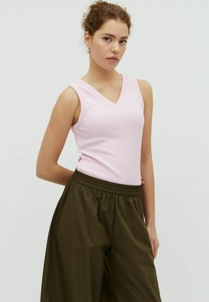 Młoda kobieta o kręconych włosach, ubrana w blado-różowy, bezrękawnik z dekoltem w serek oraz luźne, ciemnooliwkowe spodnie, stojąca z jedną ręką za plecami.