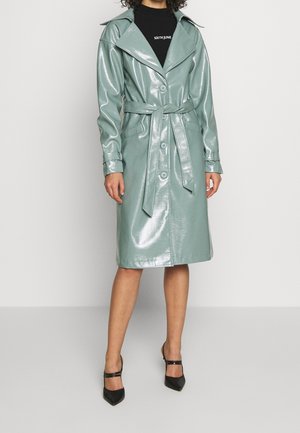 Kvinde iført en skinnende mintgrøn trenchcoat med bælte over en sort rullekrave og sorte spidse hæle, stående foran en ensfarvet baggrund.
