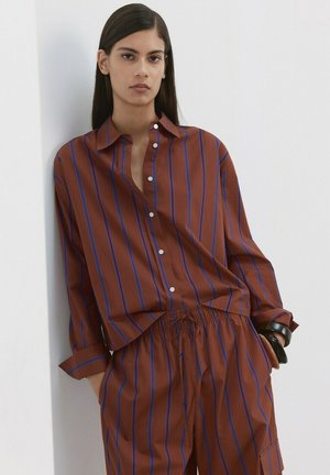 Mladý člověk s dlouhými tmavými vlasy, oblečený v hnědo-modře pruhované košili na knoflíky a odpovídajících kalhotách, stojící před bílou zdí.